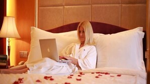 Una persona con bata blanca usando un ordenador portátil sobre una cama con ropa de cama blanca y pétalos de rosa esparcidos.