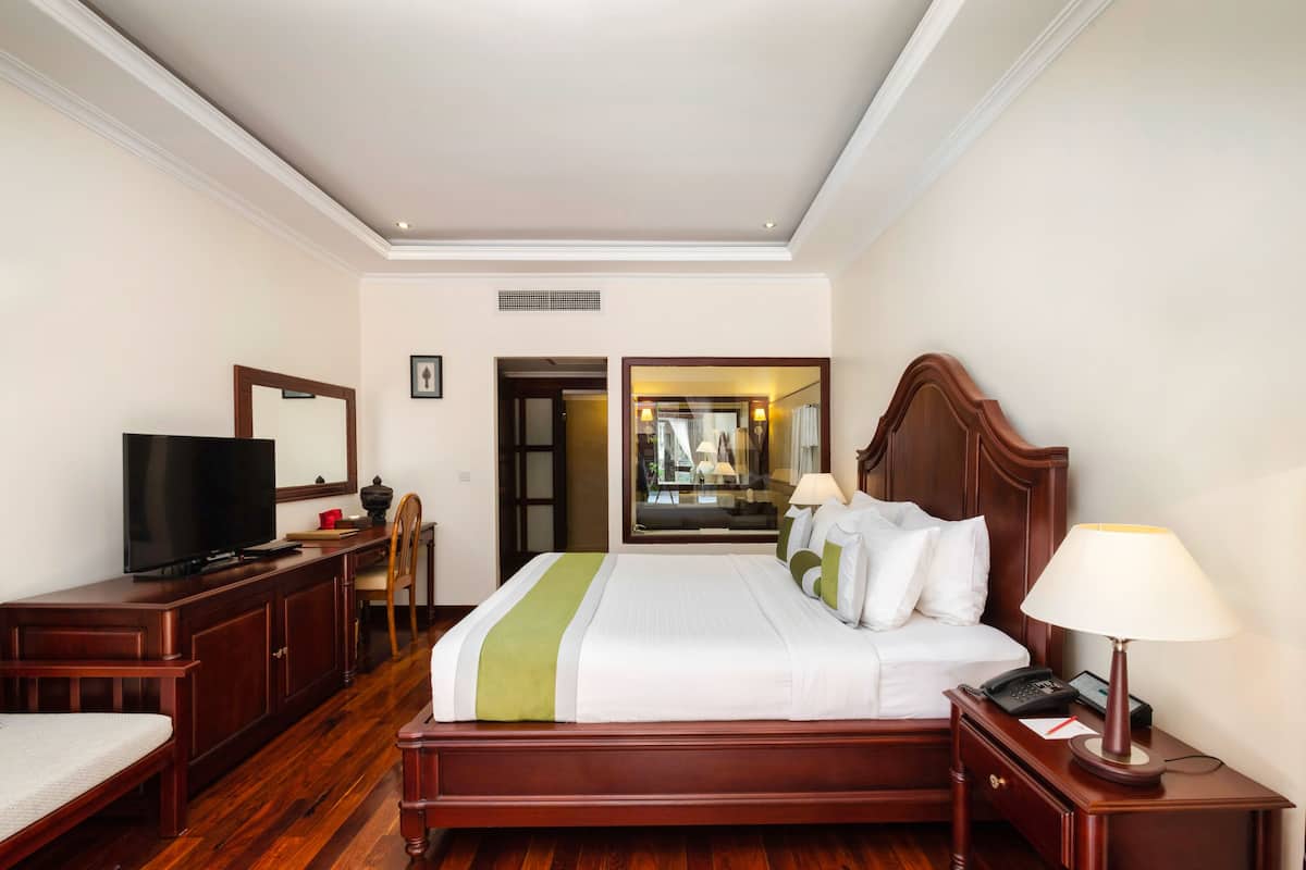 Une chambre d’hôtel avec un lit en bois, un téléviseur posé sur une commode en bois, un bureau avec une chaise et un miroir.