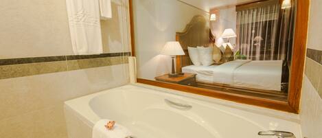 A hotel bathroom with a large bathtub, a mirror reflecting the room, a bed with white linens, and a nightstand with a lamp.