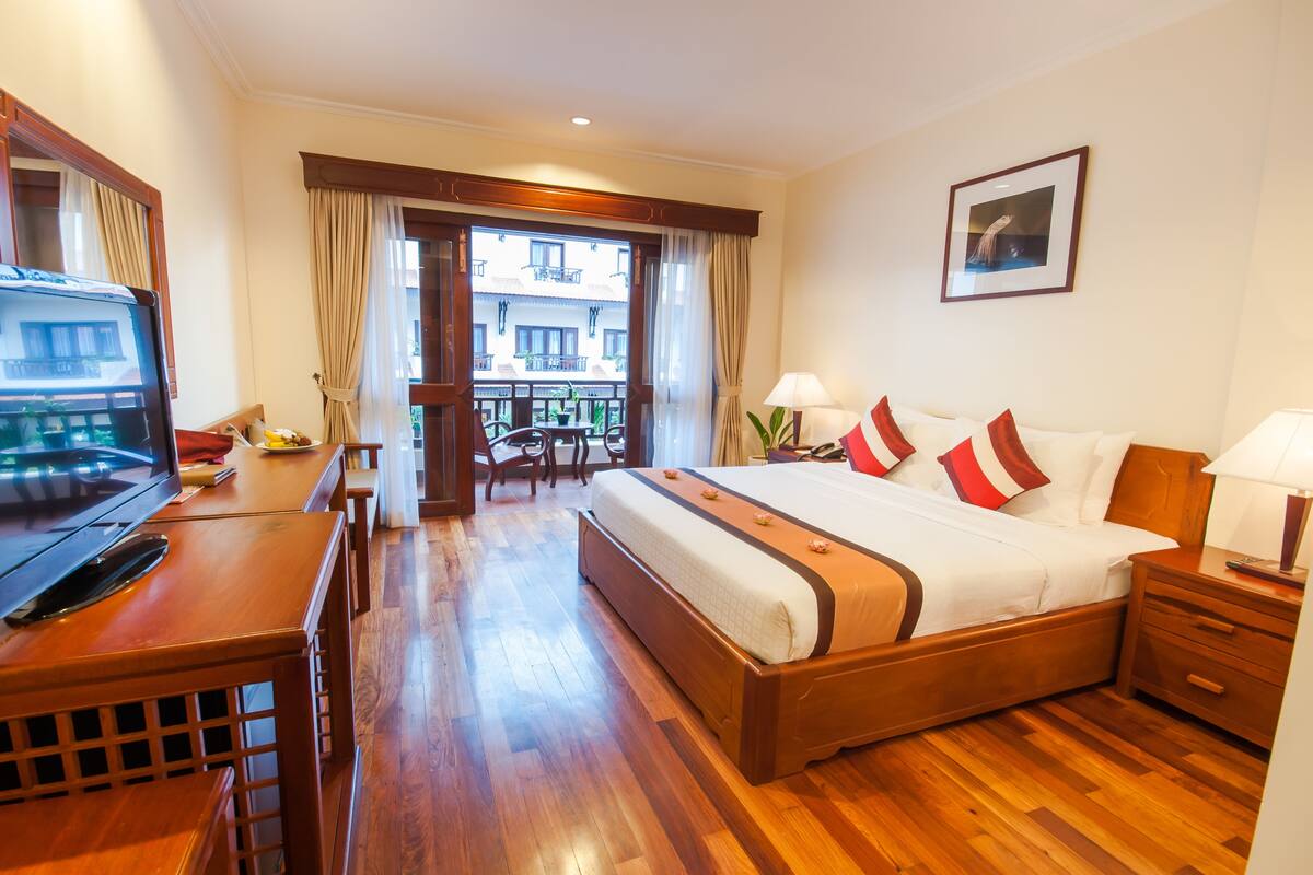 Une chambre d’hôtel avec un lit en bois, un bureau avec une télévision, un balcon avec des rideaux et un tableau encadré au mur.
