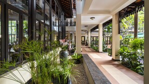 Un corredor interior amplio con suelo de baldosas, plantas en macetas y vistas al exterior a través de grandes ventanales.