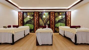 Una habitación con tres camas, cada una con una colcha blanca y una mesita auxiliar. El suelo es de madera y hay un mural grande en la pared con motivos tropicales.