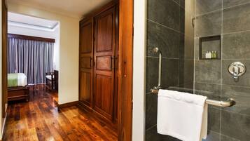 A hotel room with a wooden floor, a large wardrobe, a shower with a glass door, and a small shelf with toiletries.