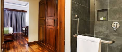 A hotel room with a wooden floor, a large wardrobe, a shower with a glass door, and a small shelf with toiletries.