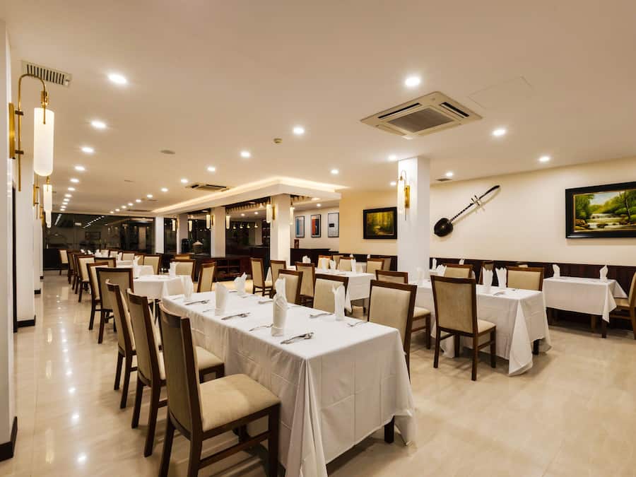 A restaurant with multiple tables and chairs, white tablecloths, and ceiling lights.