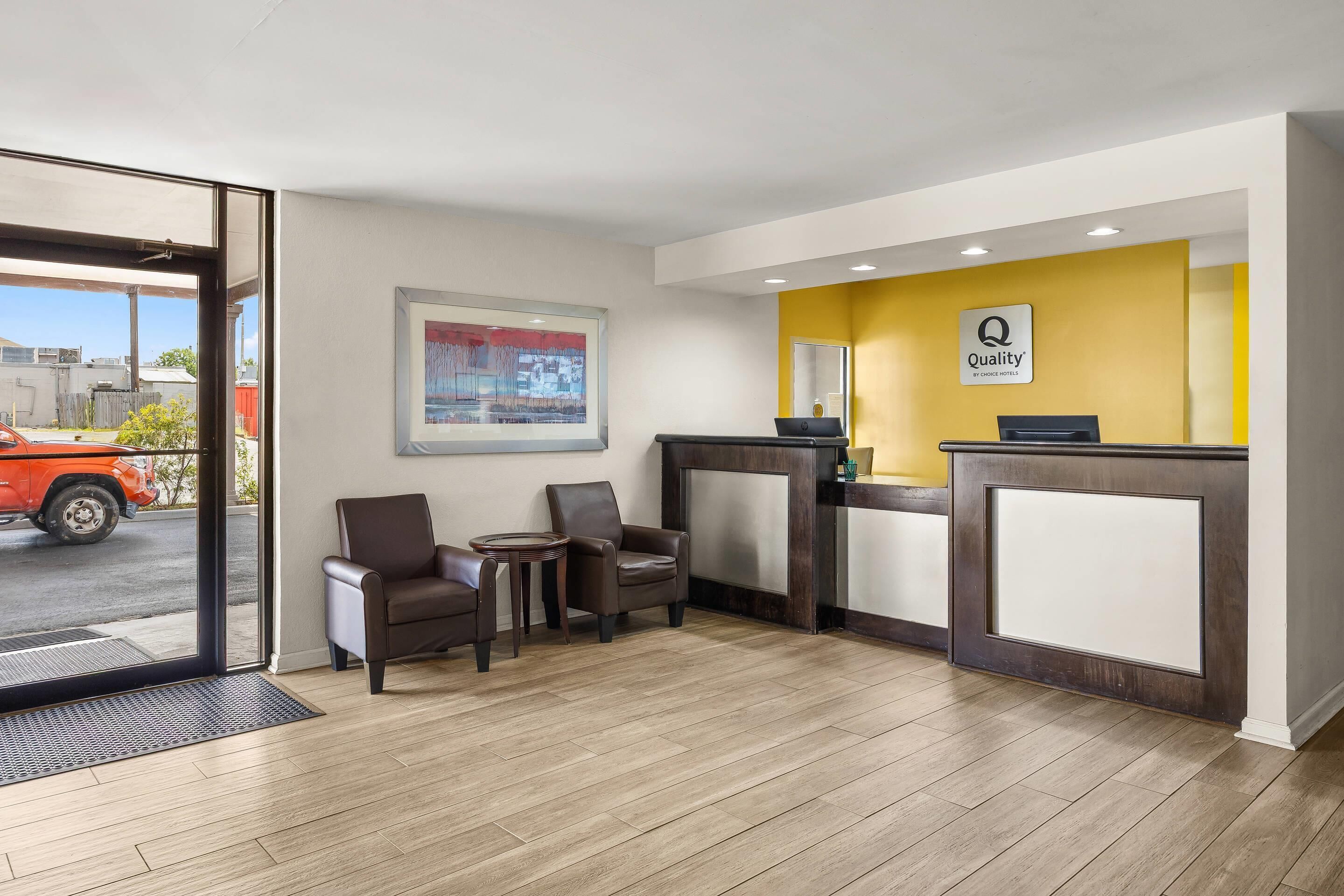 Ein Hotel-Rezeptionsbereich mit Empfangstresen, zwei Sesseln, einem kleinen Tisch und einem gerahmten Kunstwerk an der Wand.