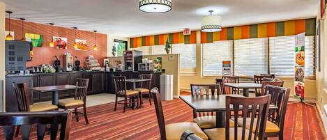 A hotel dining area with multiple tables and chairs, a kitchen in the background, and a striped wall design.
