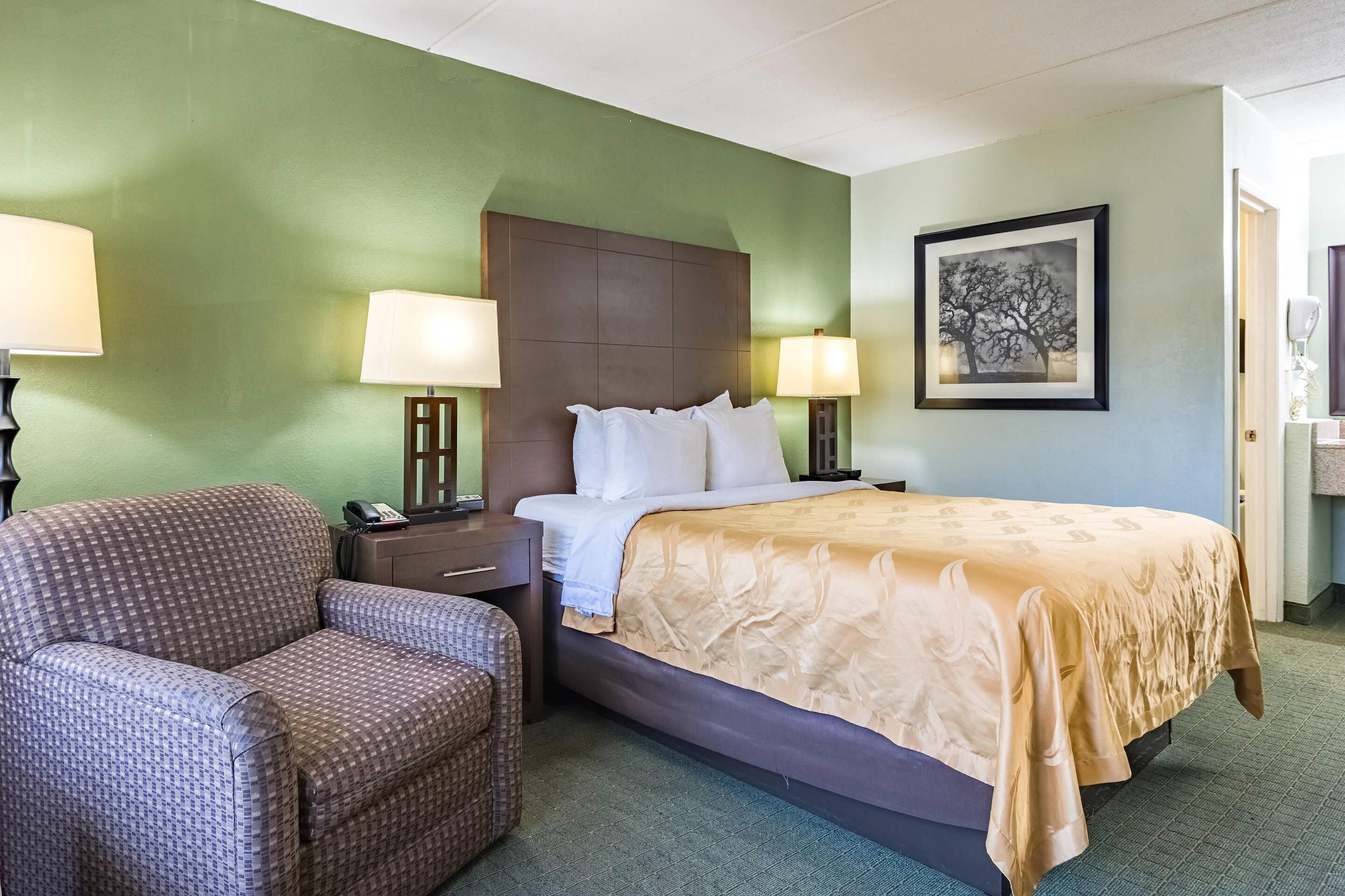 Habitación de hotel con cama, dos lámparas de noche, una silla, una mesita de noche y un cuadro en la pared.