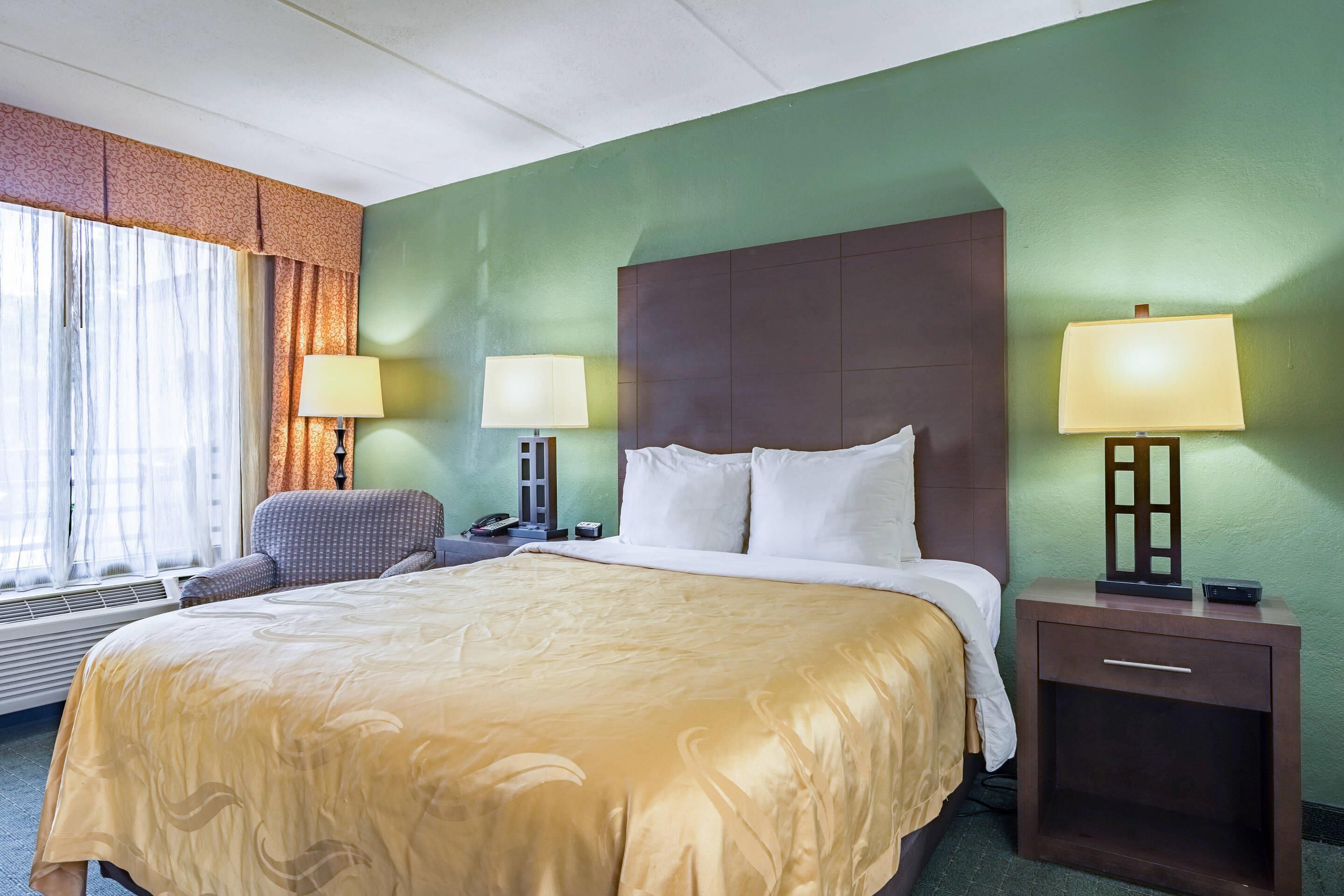 A hotel room with a large bed, two bedside lamps, a chair, a desk with a telephone, and a window with curtains.