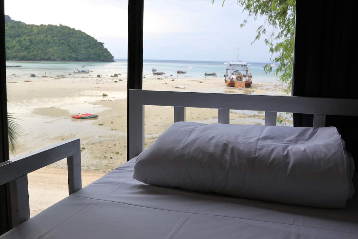 Un lit avec un oreiller blanc et une vue sur une plage où l’on aperçoit des bateaux et un camion.