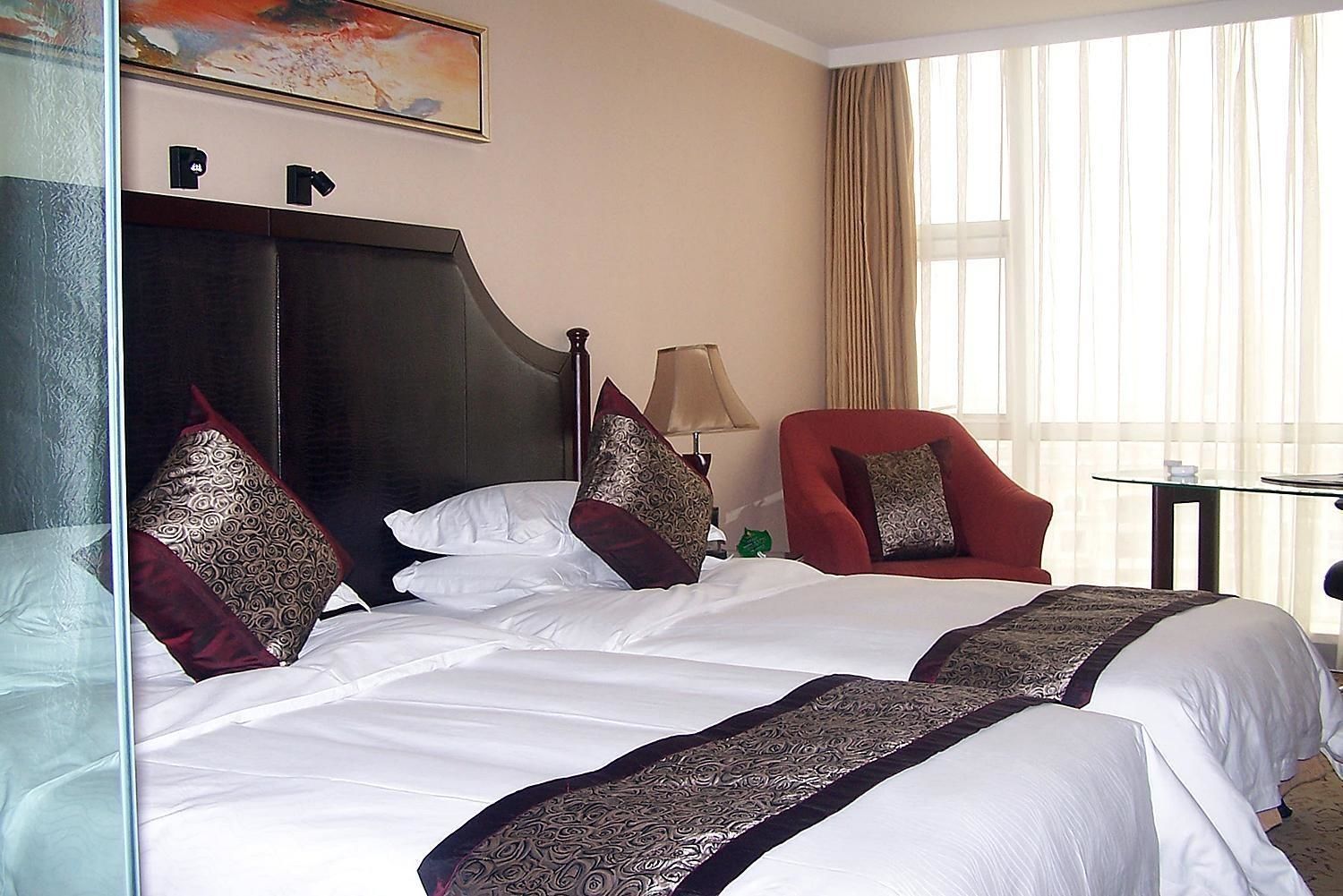 A hotel room with two beds, a red armchair, a small table, and a large window with curtains.