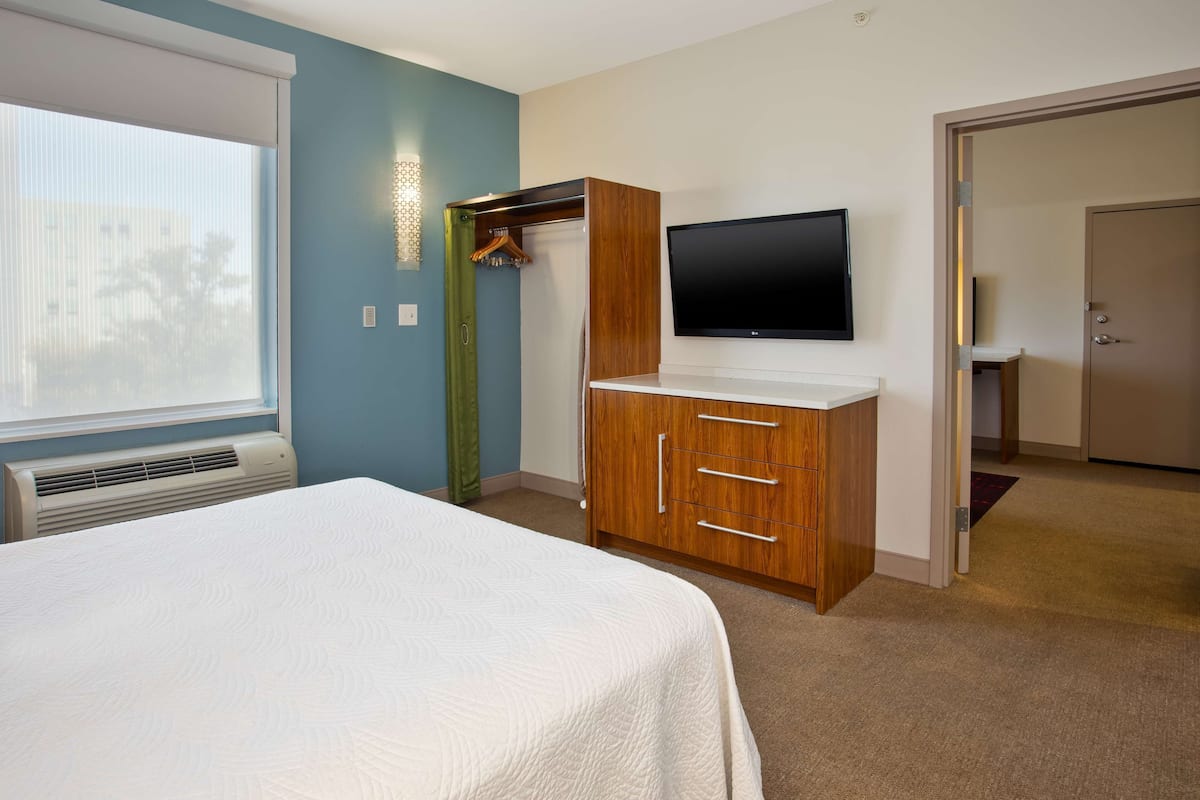 A hotel room with a bed, a TV mounted on the wall, a wooden wardrobe, and a window with blinds.