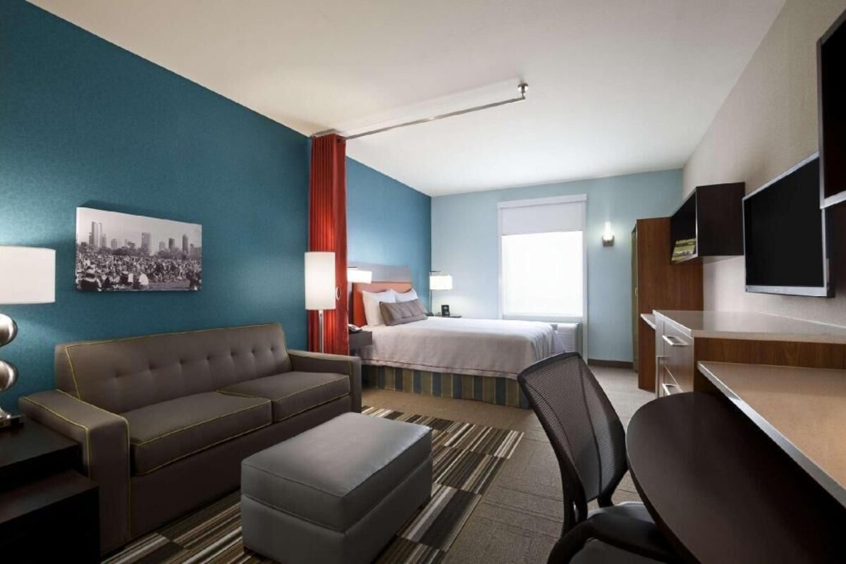 A hotel room with a bed, a sofa, a desk, a chair, a TV, and a cityscape painting on the wall.