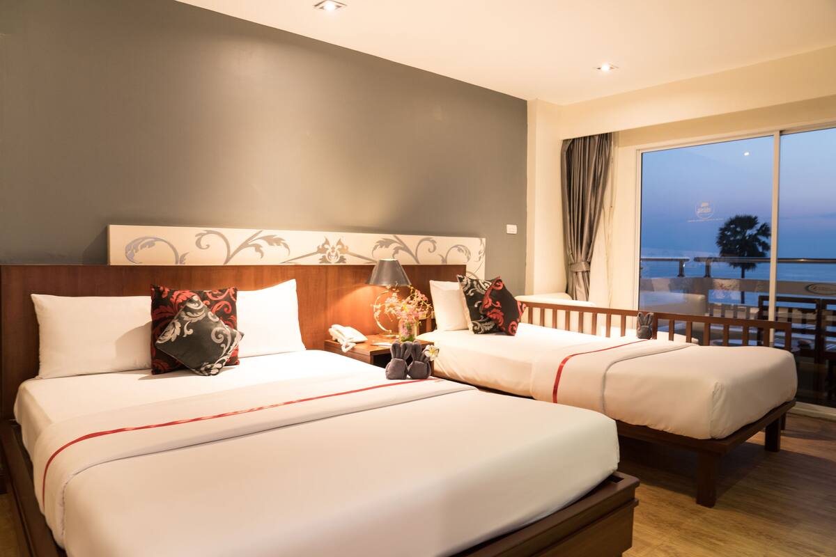 A hotel room with two beds, a nightstand, a lamp, and a view of the sea.
