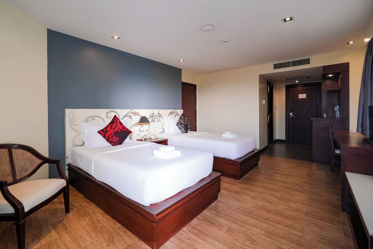 A hotel room with two beds, a wooden chair, a desk, and a kitchenette.