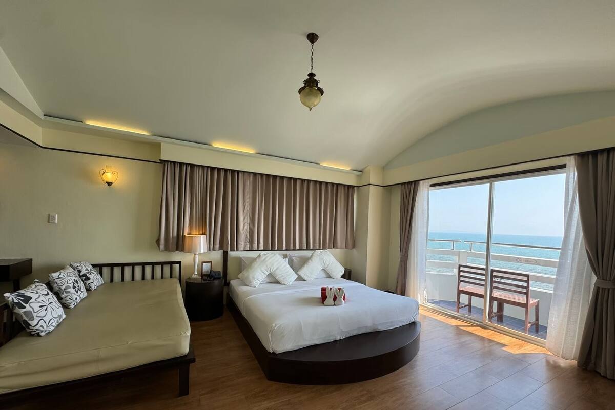 A hotel room with a bed, a sofa, a nightstand, a lamp, and a view of the ocean through a large window.