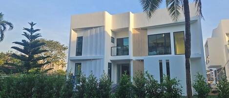 A modern two-story building with large windows and a flat roof, surrounded by greenery and palm trees.
