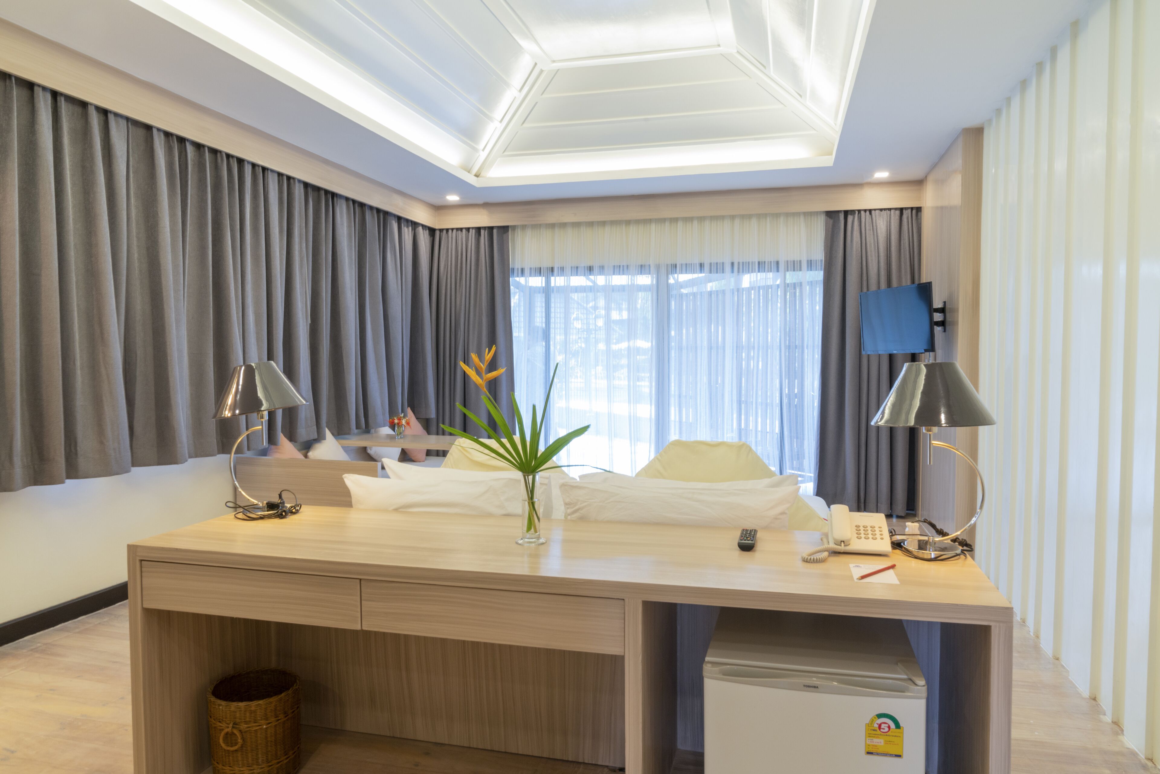 A hotel room with a wooden desk, two lamps, a small plant, a TV, and a minibar.