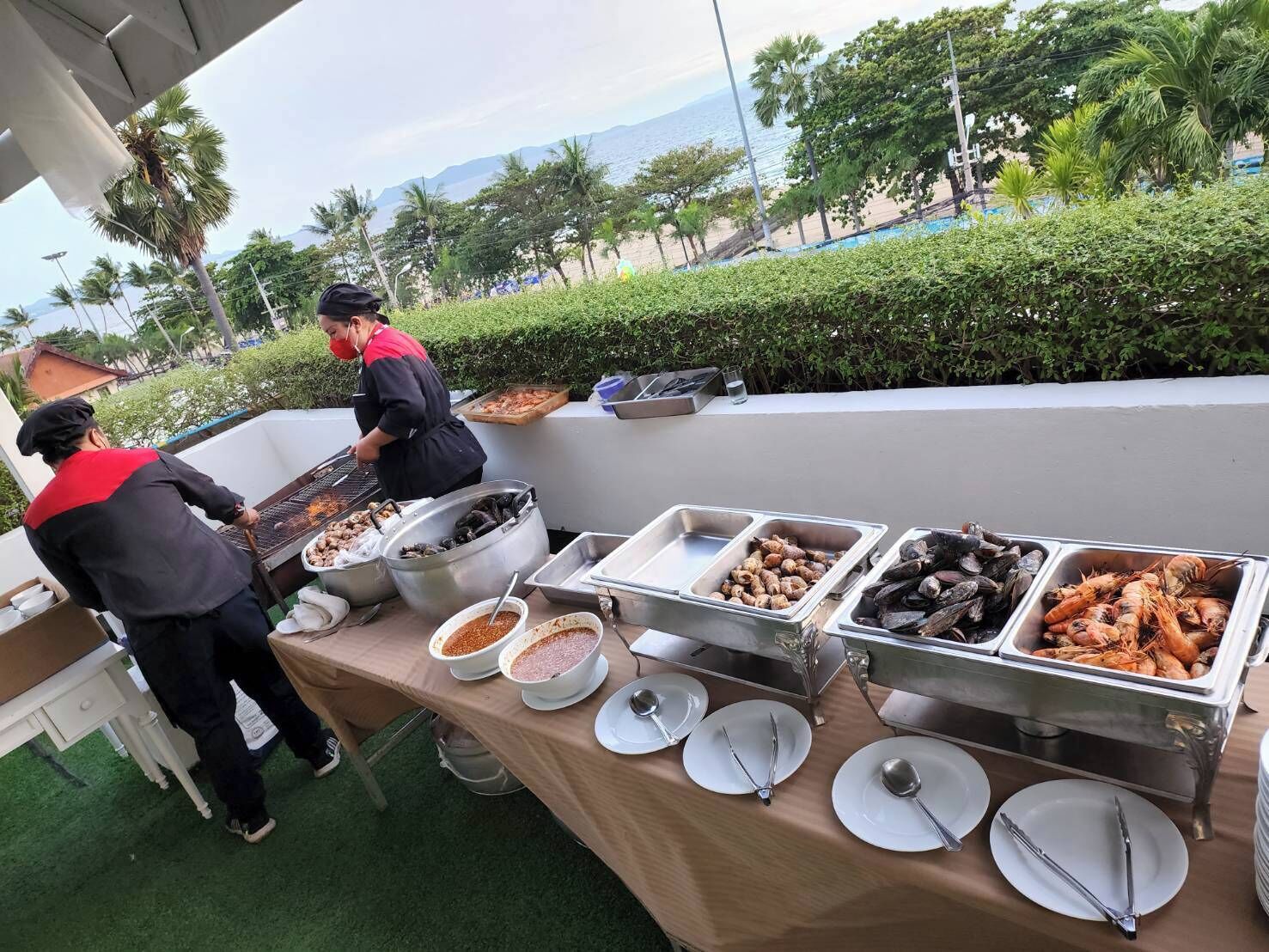 Ein Buffet mit Meeresfrüchten und Soßen, eine Person grillt, und ein schöner Blick auf Strand und Berge.