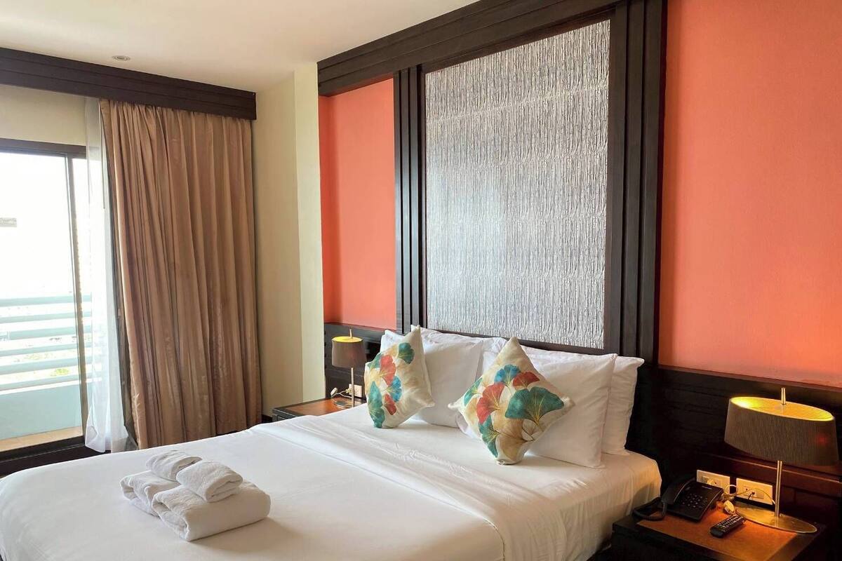 A hotel room with a bed, pillows, a nightstand, a lamp, and a view of the outside.