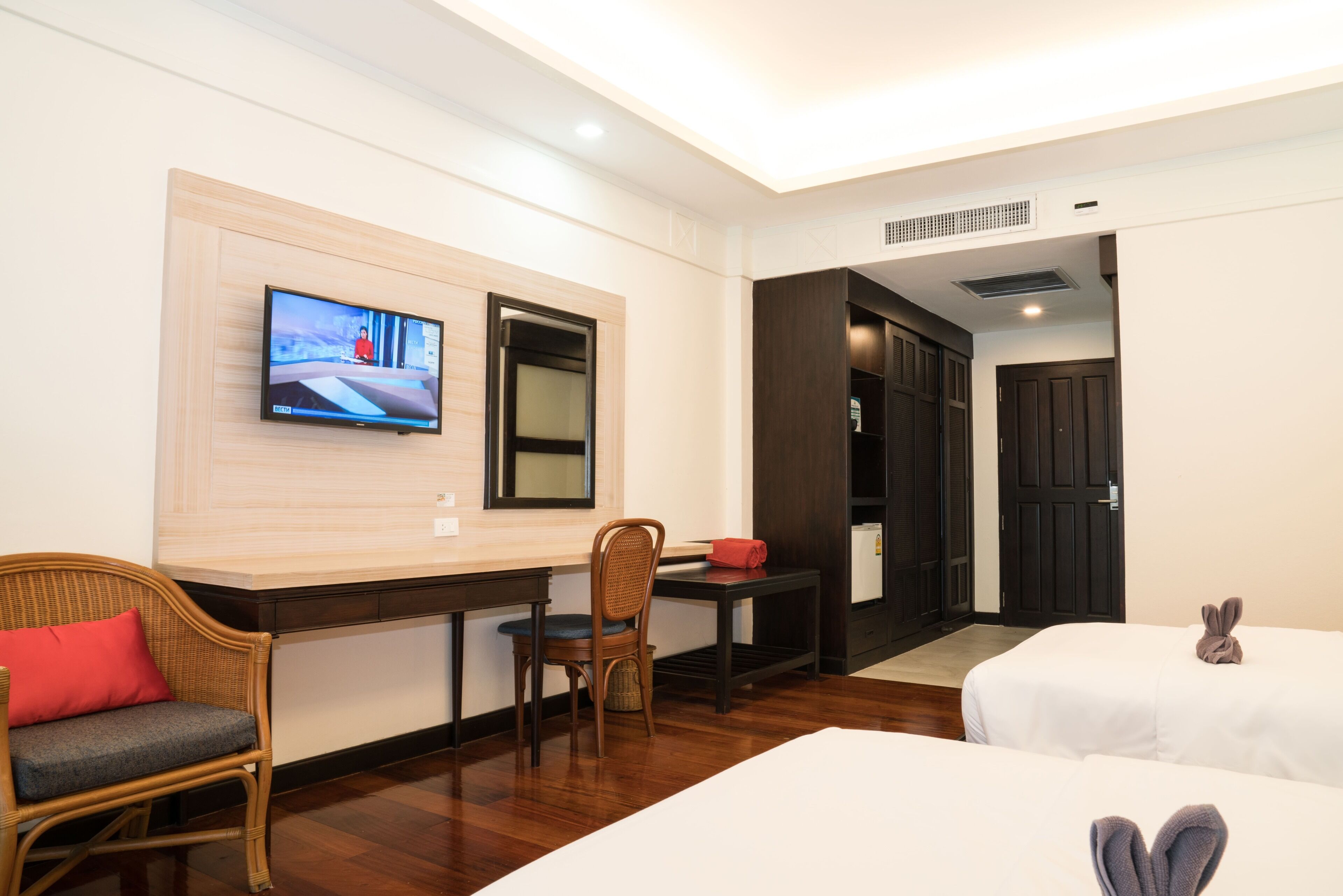 A hotel room with a TV mounted on the wall, a dining table with chairs, two beds with towels, and a door leading to another room.