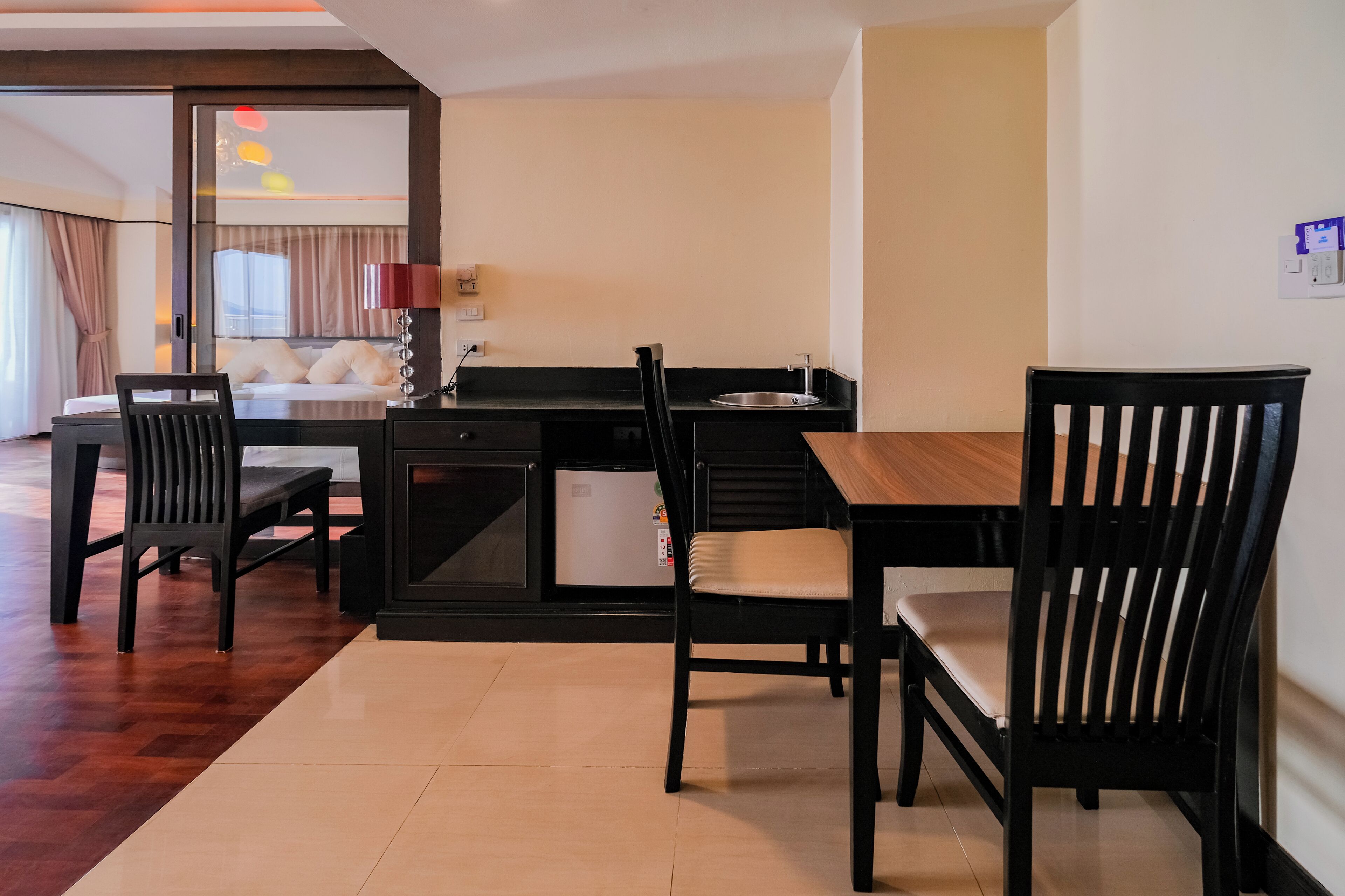 A hotel room with a bed, a desk, a chair, a small kitchen area, and a dining table.