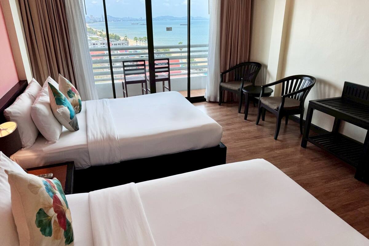 A hotel room with two beds, a desk, and a chair. There is a view of the sea and buildings through the window.