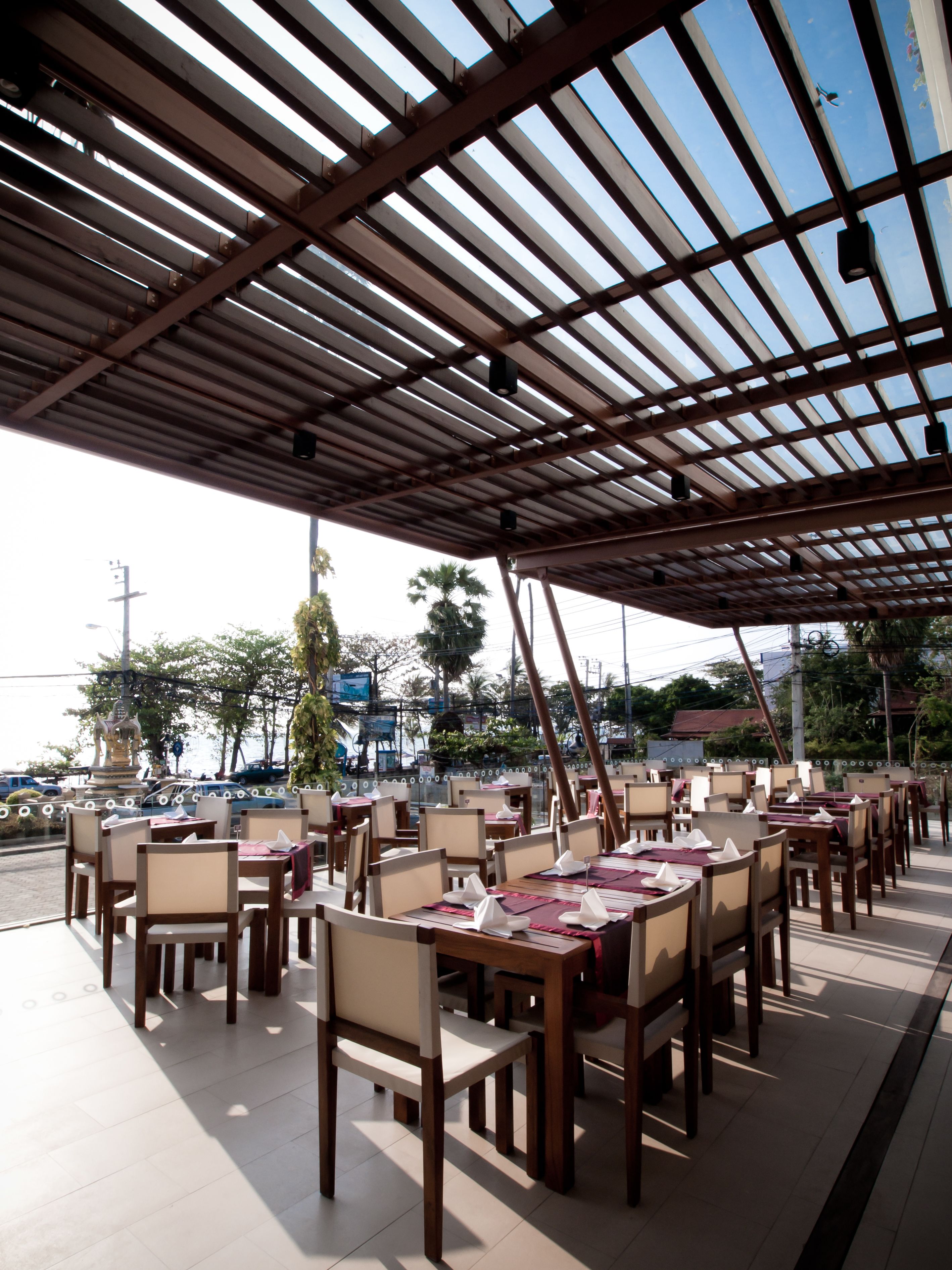 An outdoor restaurant with a covered patio, wooden tables and chairs, and a view of the street and palm trees.