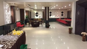 A spacious hall with a long table set for a buffet, surrounded by red chairs, and a row of tables with red tablecloths in the background. - Hotel Namaskar Residency (Amritsar)