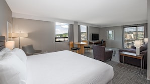 A hotel room with a large bed, a desk, a chair, a sofa, a TV, and large windows with a city view.