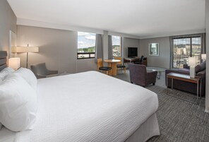 Una habitación de hotel con una cama grande, un escritorio, una silla, un sofá, un televisor y ventanas amplias con vista a la ciudad.