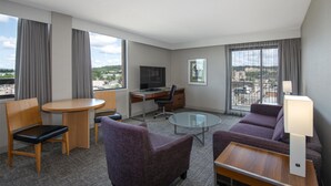 Quarto de hotel com uma escrivaninha, dois sofás roxos, uma mesa de vidro redonda, uma televisão e vista para a cidade.