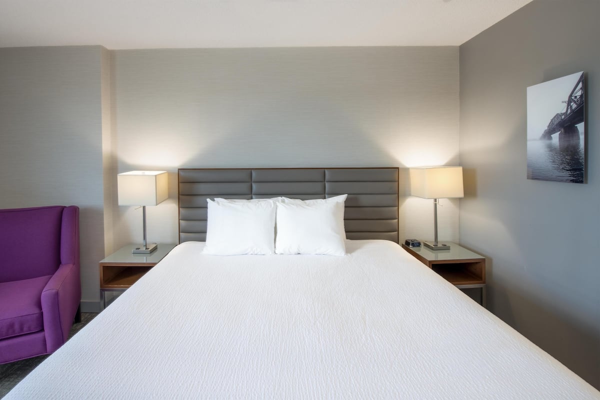 A hotel room with a large bed, two bedside lamps, a purple armchair, and a framed picture on the wall.