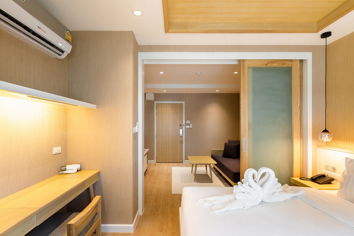 Een hotelkamer met een bed, bureau, stoel, nachtkastje, spiegel, een luchtverkoeler aan de muur en een deur naar een andere kamer.