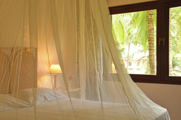Una cama con dosel, una lámpara de noche y una ventana con vista a palmeras.
