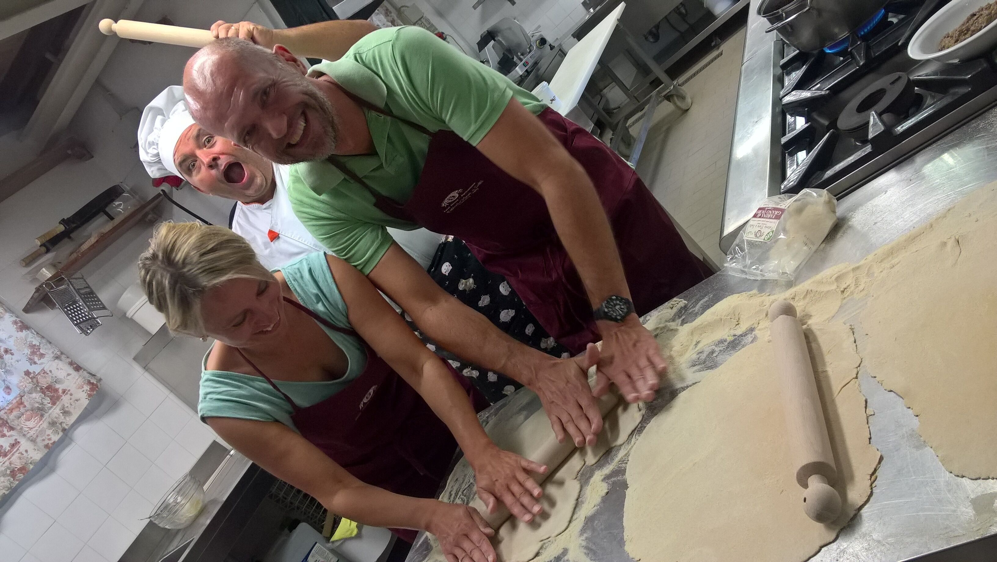 Duas pessoas estendem massa em uma bancada de cozinha, usando um rolo de massa e uma folha grande de massa.