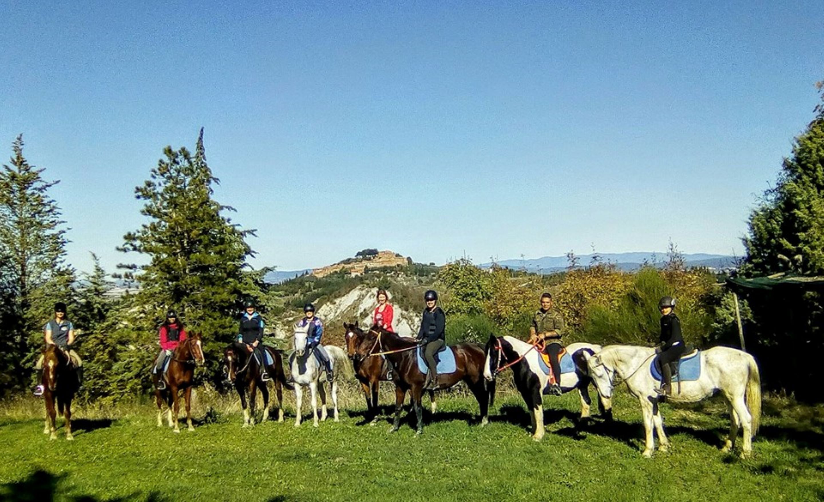 A group of people on horseback in a grassy field with trees and a hill in the background.