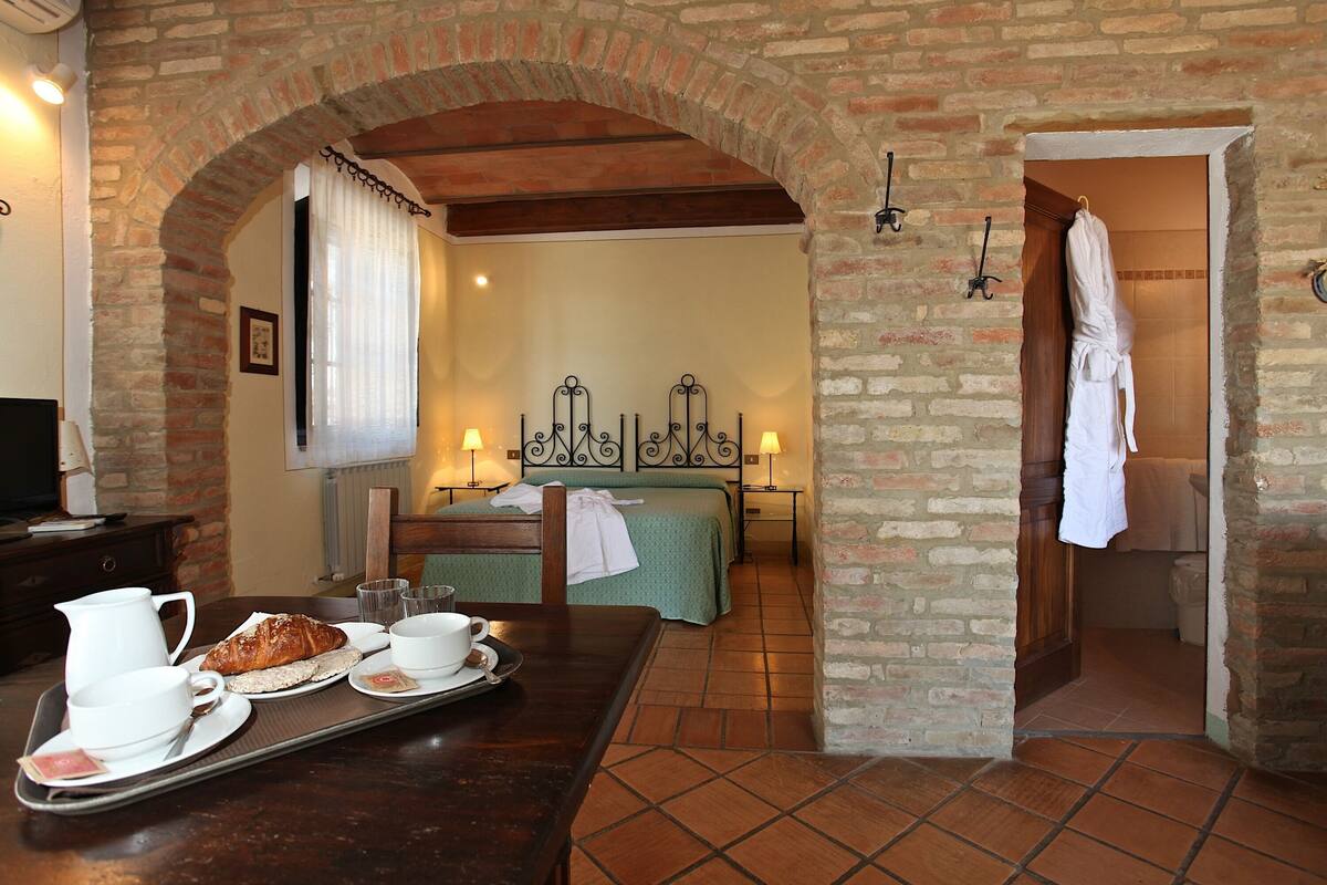 Une chambre d’hôtel avec un mur en briques, un lit recouvert d’une courtepointe verte, une table en bois sur laquelle sont disposés des articles pour le petit-déjeuner, et une salle de bain visible à travers une arche.