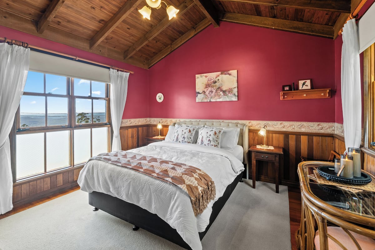 Une chambre avec un grand lit, un plafond en bois, une fenêtre avec des rideaux, une table de chevet avec une lampe et une horloge murale.