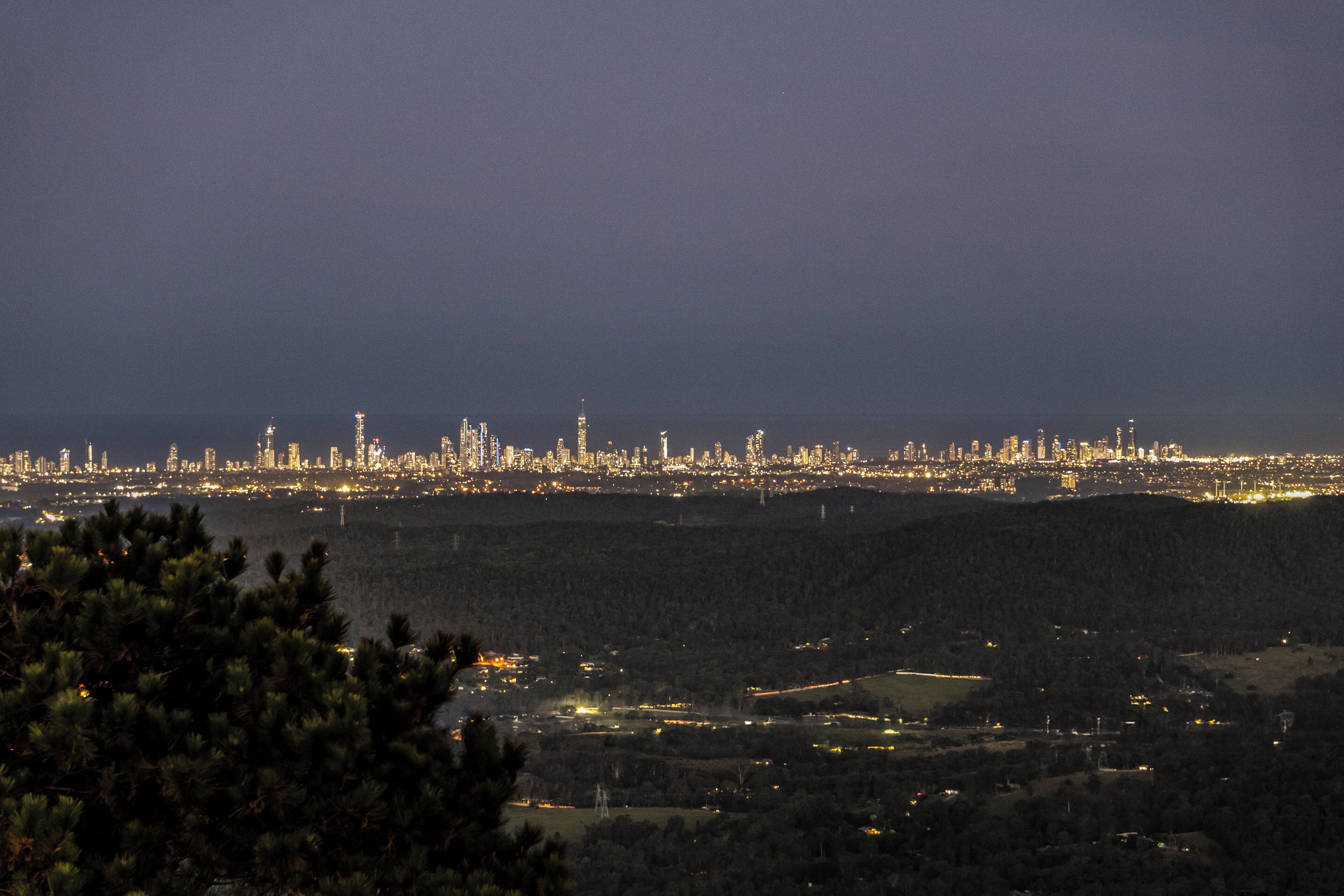 A nighttime cityscape with a distant view of a city skyline, a forested area in the foreground, and a body of water.