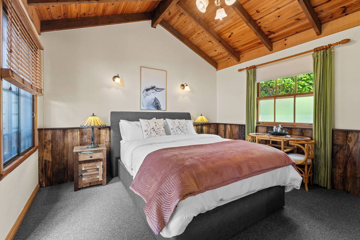 Une chambre à coucher avec un lit, des tables de chevet, une fenêtre avec des rideaux, un plafond en bois et un tableau accroché au mur.