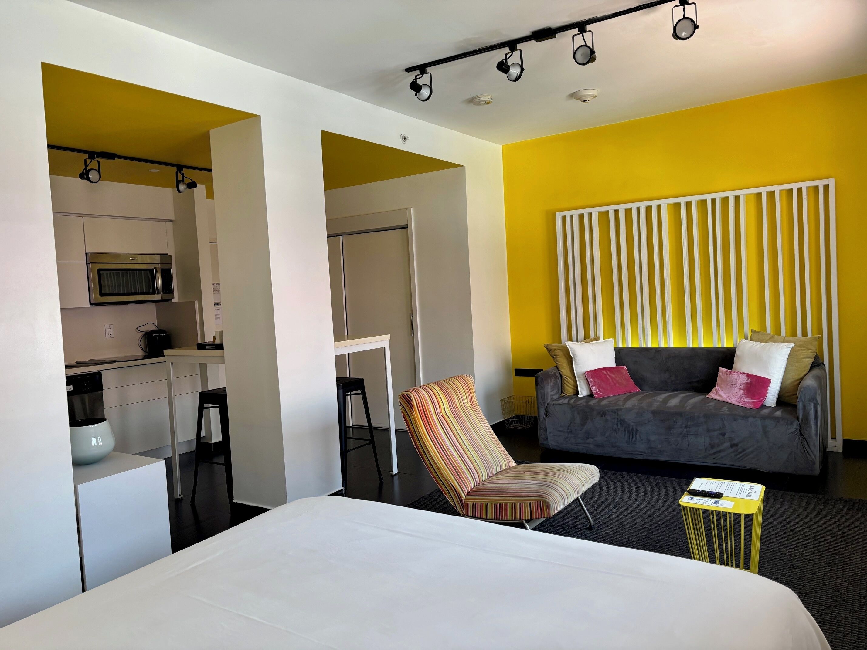 Une chambre d’hôtel moderne comprenant un lit, un canapé, une chaise à rayures, une petite table, un four à micro-ondes dans le coin cuisine et un mur à rayures jaunes.