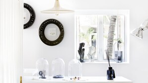 Ein Zimmer mit weißer Wand, einem Fenster mit Blick auf Palmen, einem weißen Schreibtisch mit Lampe und dekorativen Gegenständen an der Wand.