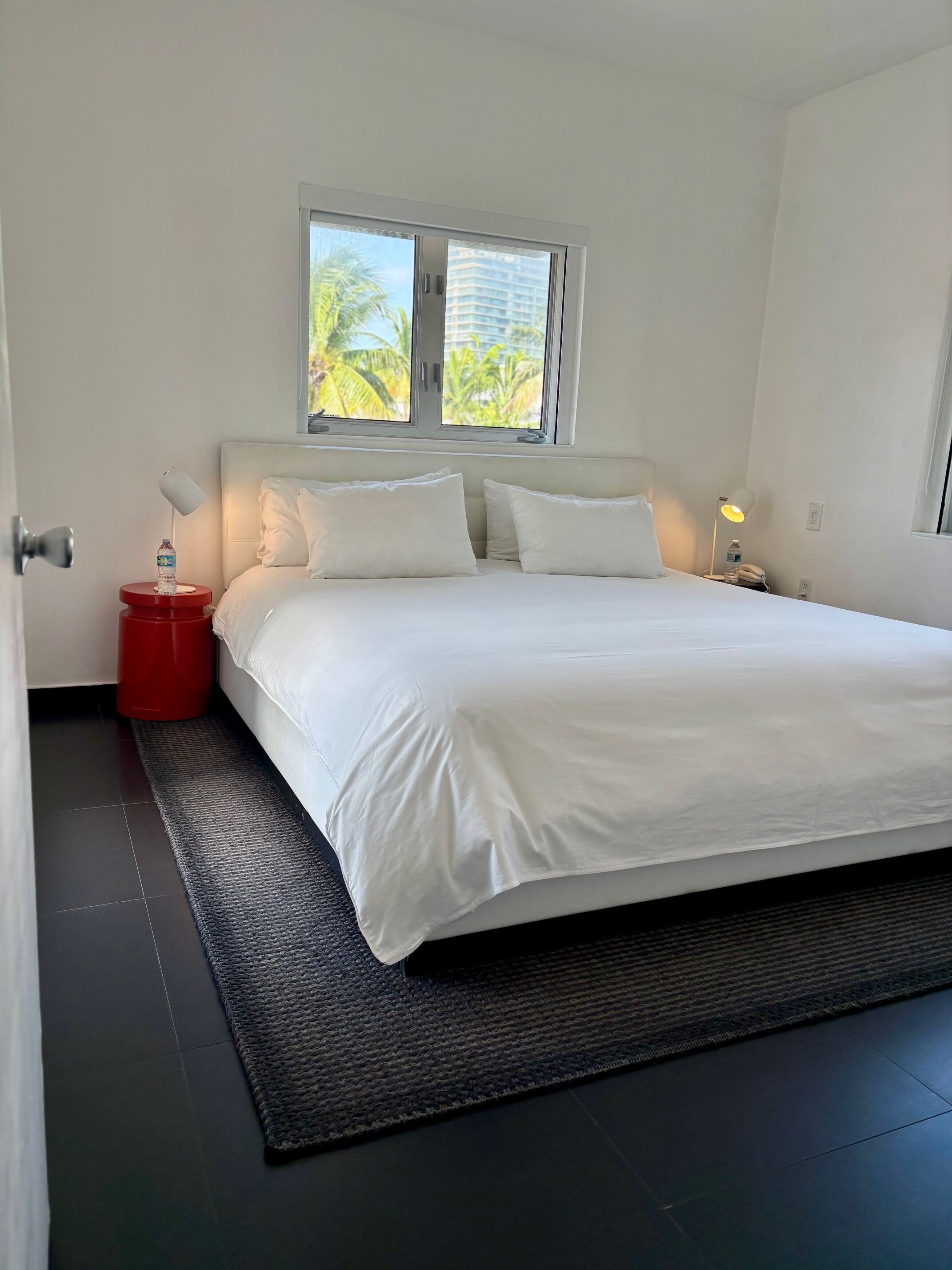 Ein Schlafzimmer mit einem Bett, einem Fenster, einem Nachttisch und einer roten Mülltonne.
