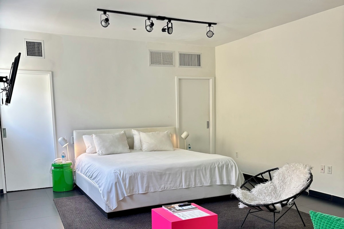A modern bedroom with a bed, a desk, a chair, and a small green trash bin.