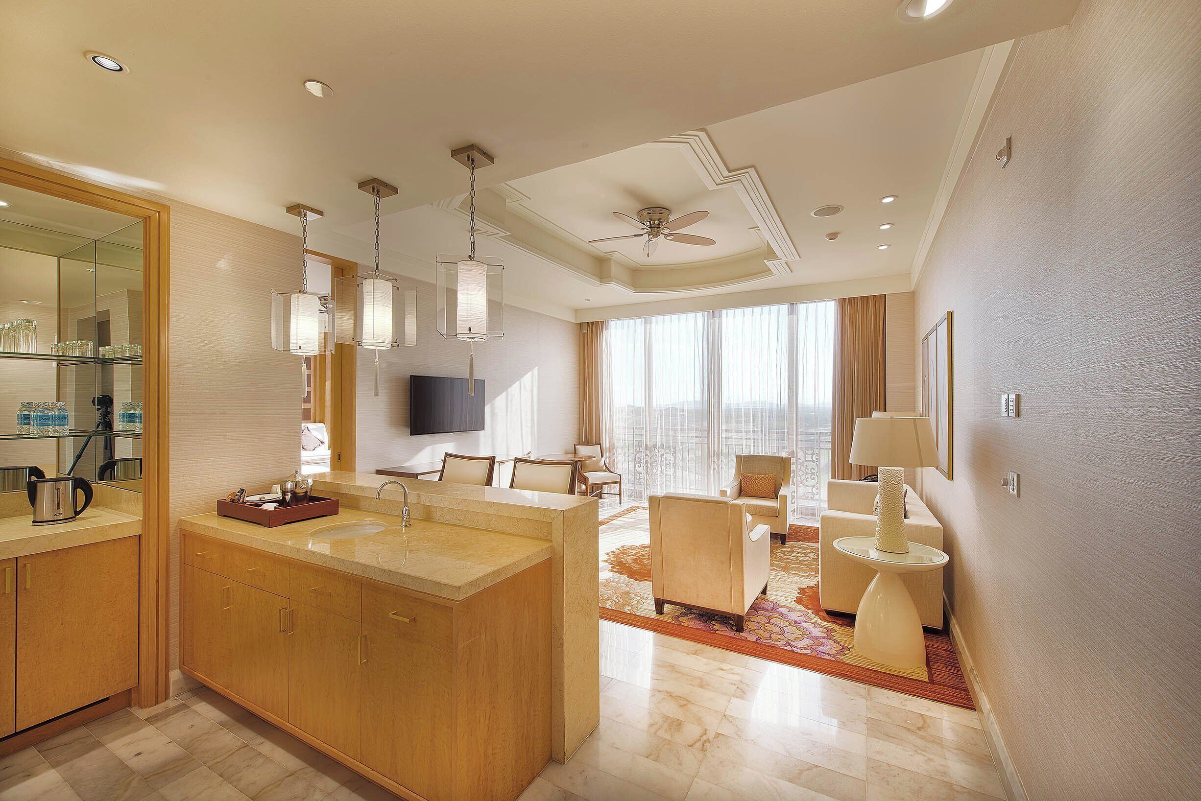 A modern hotel room with a large bathroom, a seating area with a sofa and armchairs, and a television mounted on the wall.