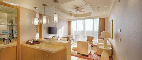A modern hotel room with a large bathroom, a seating area with a sofa and armchairs, and a television mounted on the wall.