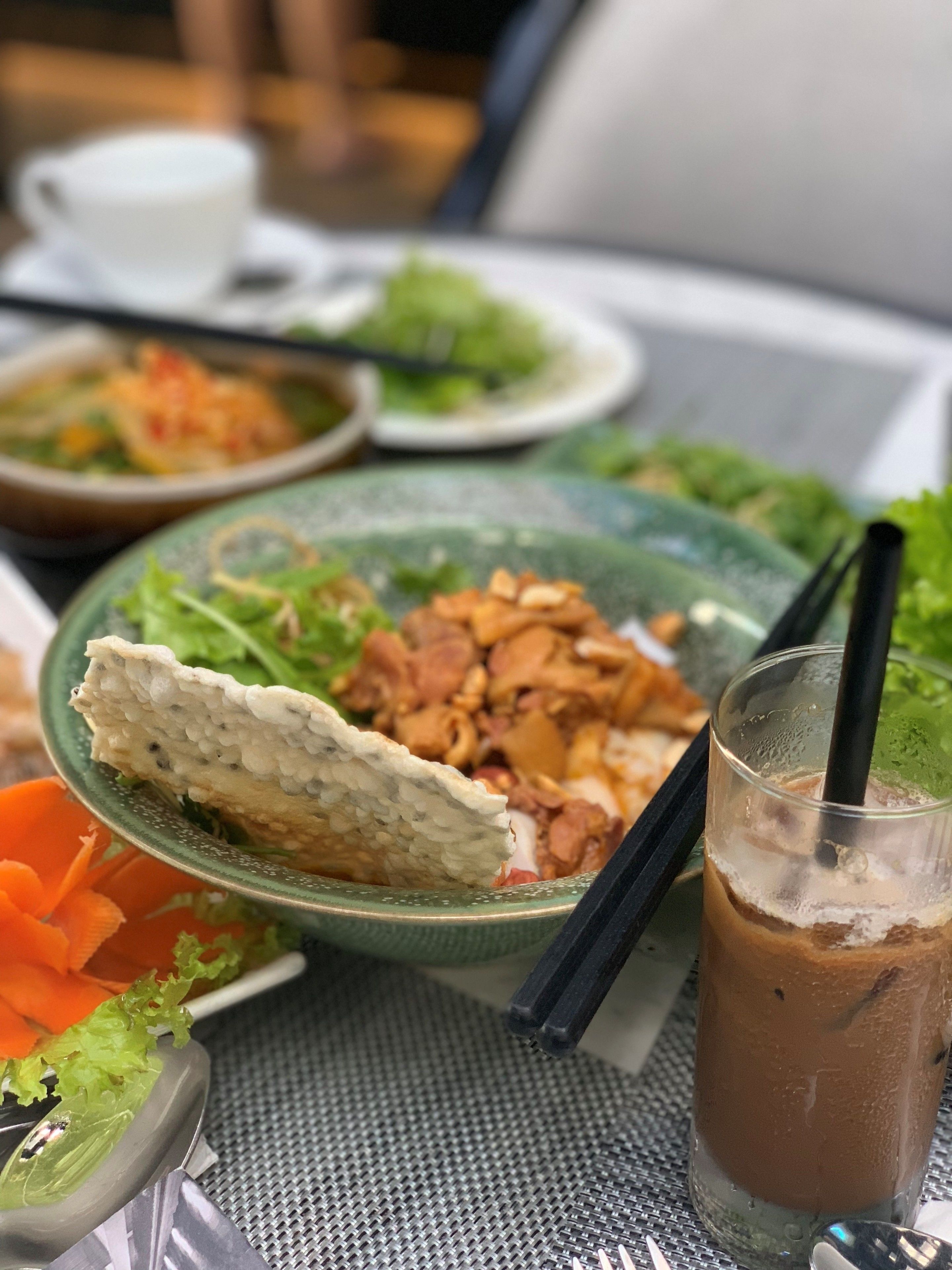 A table with a variety of dishes including a bowl of noodles, a plate of fried chicken, and a glass of iced beverage.