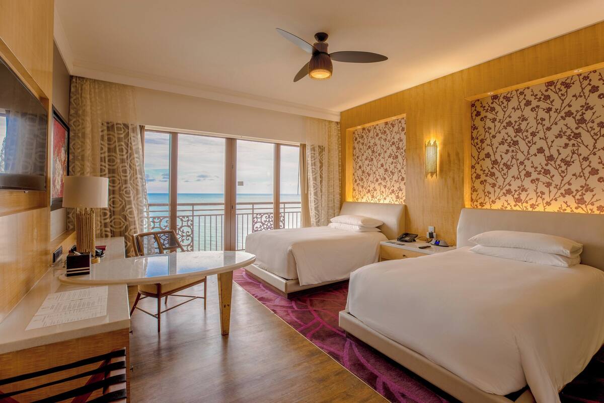 A hotel room with two beds, a desk, a chair, a table, a ceiling fan, and a view of the ocean.