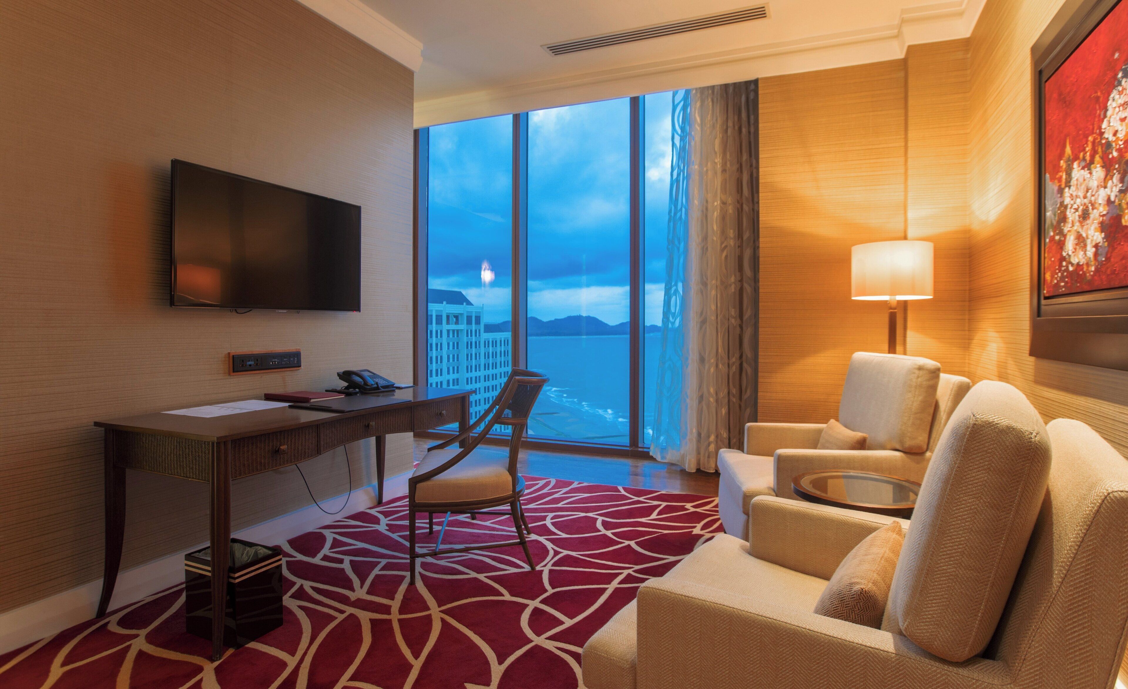 A hotel room with a large window, a desk with a flat-screen TV, a chair, a sofa, and a lamp.